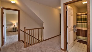 A carpeted hallway with a wooden staircase, a door leading to a bathroom with a striped shower curtain, and a lamp in the adjacent room.