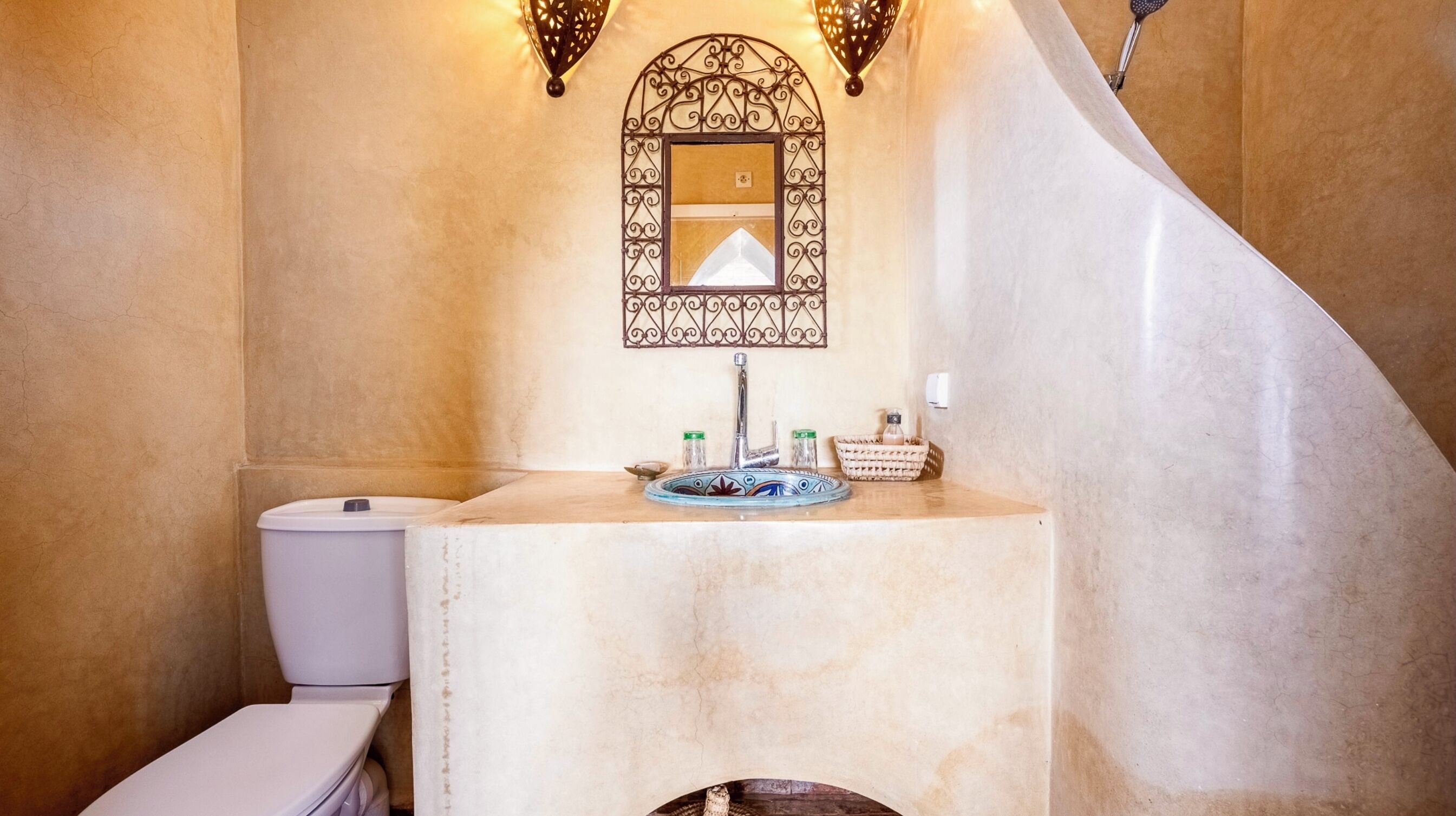 Une salle de bain avec des toilettes blanches, un lavabo doté d’un receveur décoratif et un miroir mural au motif géométrique.