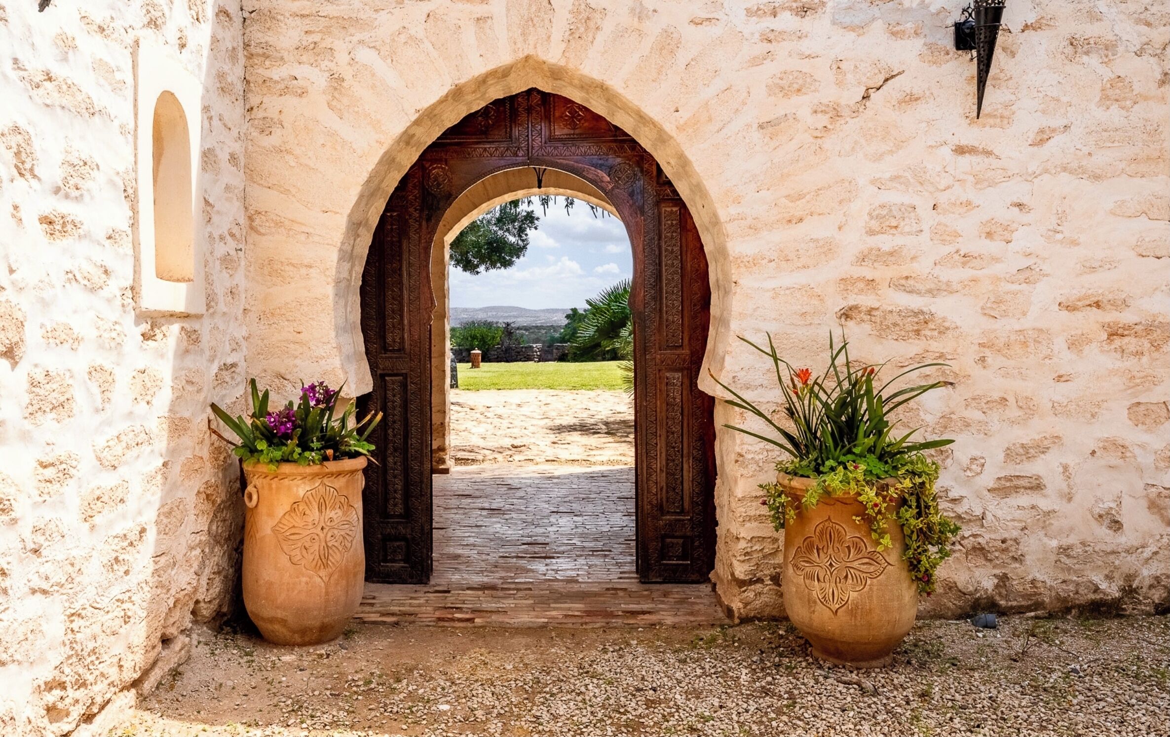 Uma porta em arco dá acesso a um pátio com um caminho de cascalho, ladeado por dois grandes vasos de terracota com plantas.