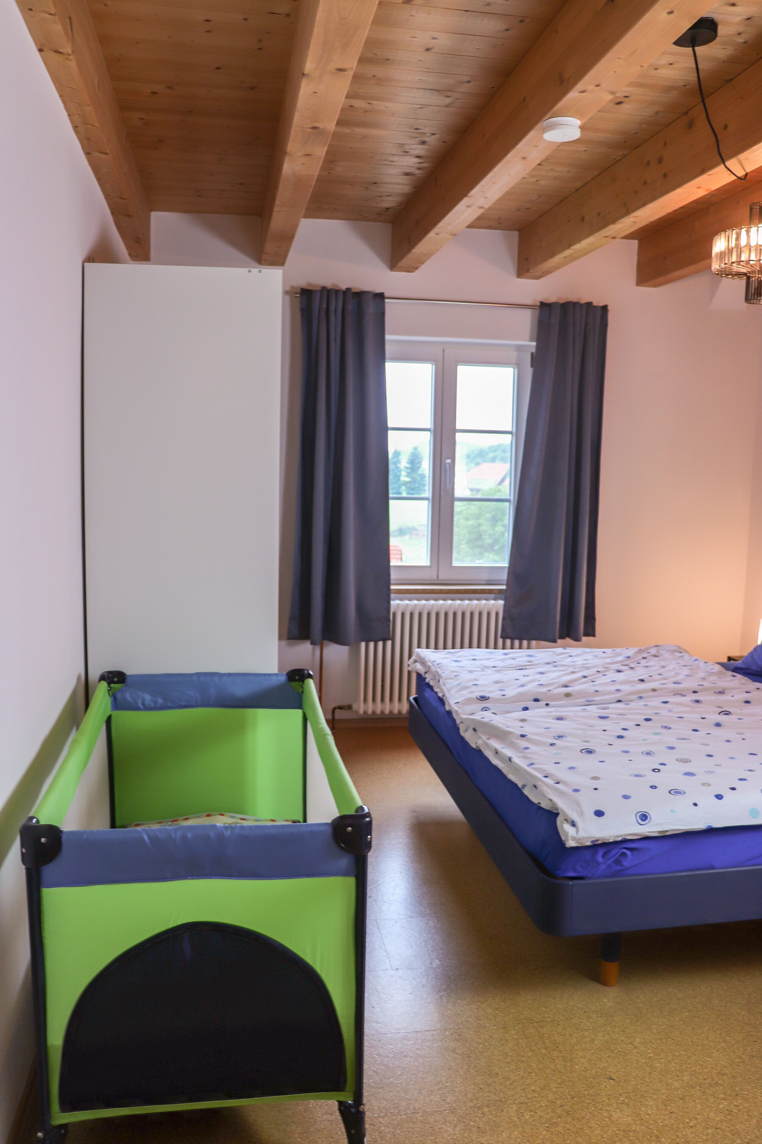Une pièce avec un lit, un parc pour enfants aux couleurs verte et bleue, une fenêtre avec des rideaux et un radiateur.