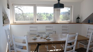A dining area with a wooden table and white chairs, large windows with a view of trees, and a hanging light fixture.