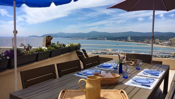 Un espace de restauration en plein air avec une table dressée pour deux personnes, offrant une vue sur la mer et les montagnes au loin.