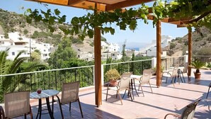 A covered outdoor patio with tables and chairs, overlooking a hillside with buildings. - Neos Ikaros (Agios Vasileios)