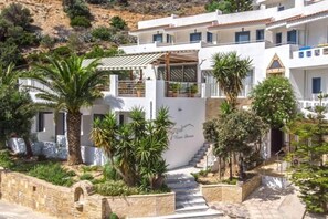 A white building with a staircase leading to a covered entrance, surrounded by palm trees and plants. - Neos Ikaros (Agios Vasileios)