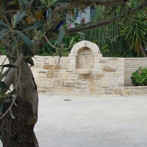 A stone wall with a small arched entrance, a tree in the foreground, and a building in the background. - Neos Ikaros (Agios Vasileios)