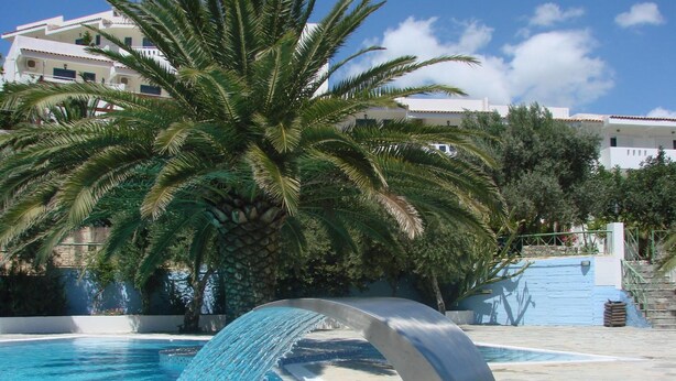 A swimming pool with a water feature, surrounded by palm trees and buildings. - Neos Ikaros (Agios Vasileios)