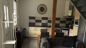 A modern living room with a grey sofa, a patterned ottoman, and a dining area with a wooden table and chairs. A kitchen with a checkered backsplash and modern appliances is visible in the background.
