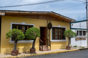 A yellow building with a wooden roof, a staircase, and three potted trees in front.