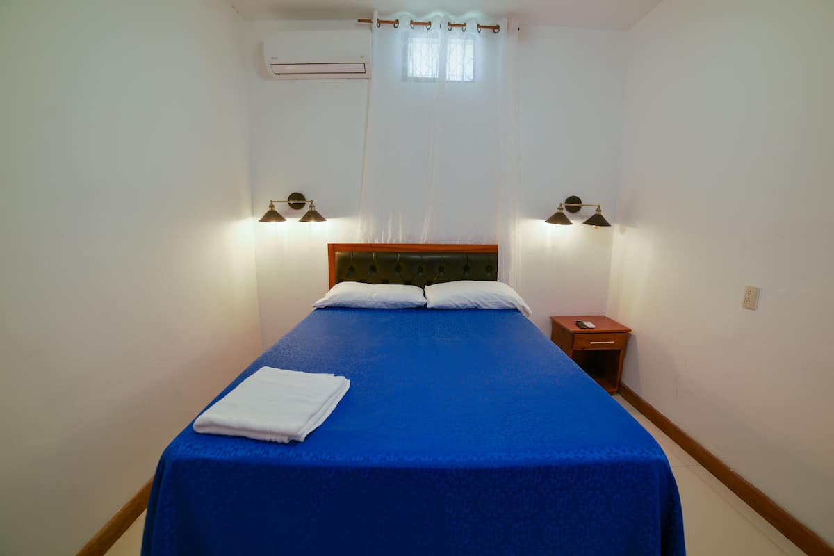 A neatly made bed with a blue bedspread and white pillows, two bedside lamps, and a small wooden nightstand with a remote control.