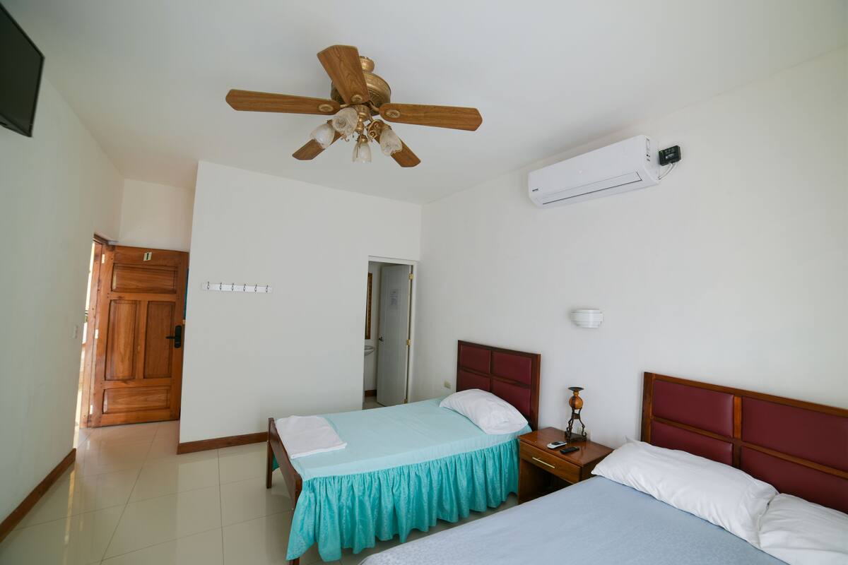 A bedroom with two beds, a ceiling fan, an air conditioner, a wooden door, and a tiled floor.
