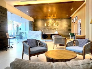 A hotel lobby with a reception desk, seating area with chairs and a round table, and a large window providing a view of the outdoors.