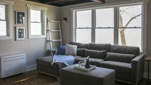 Una sala de estar con un sofá gris, una mesita y una escalera apoyada en la pared. Hay cuadros enmarcados en la pared y una ventana con vistas al exterior.