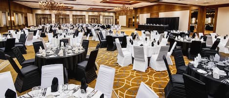 Een ruime balzaal met ronde tafels gedekt voor een diner, zwarte stoelen en witte tafelkleden.