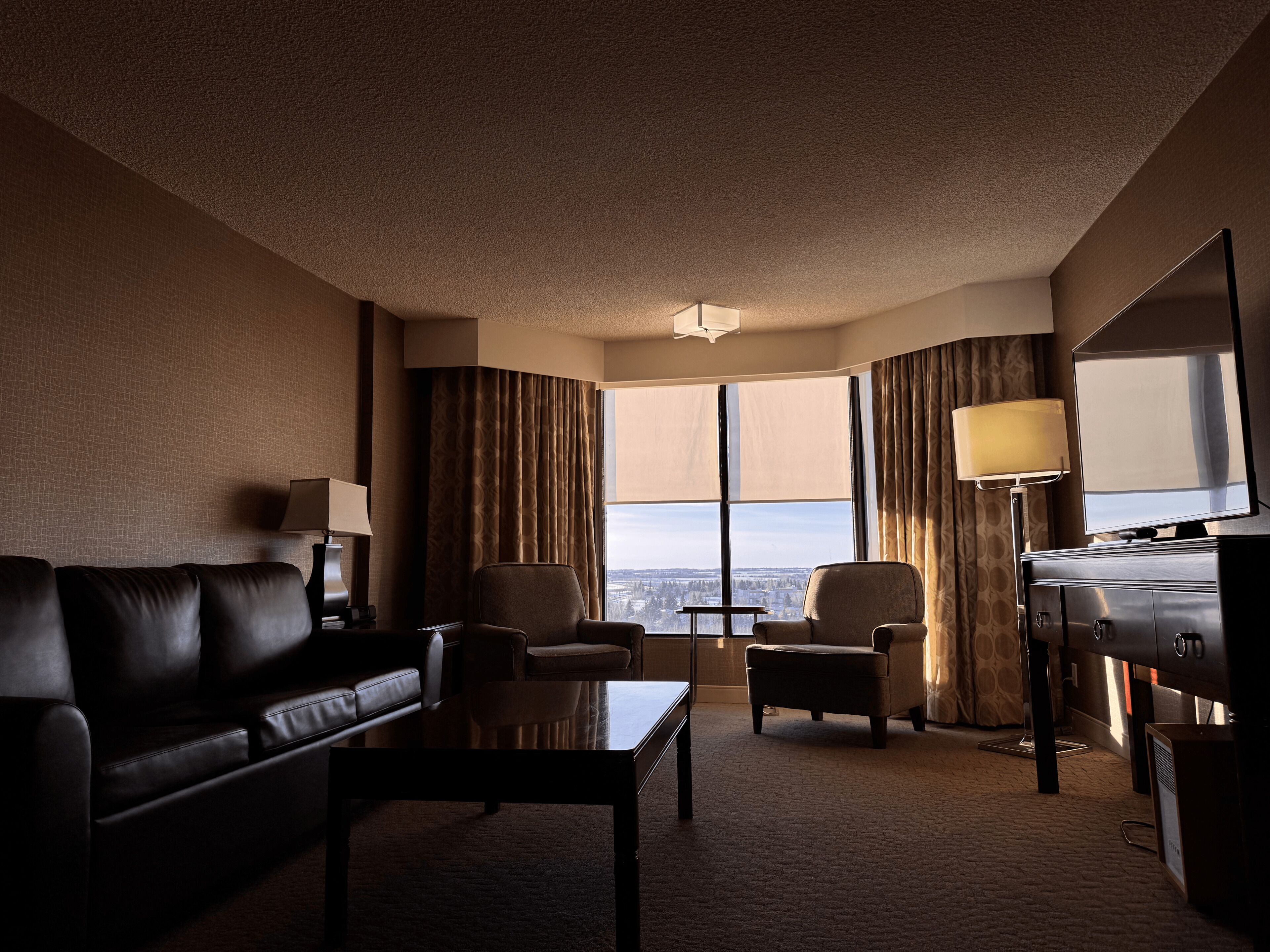 A hotel room with a leather sofa, two armchairs, a coffee table, a TV, and a large window with curtains.
