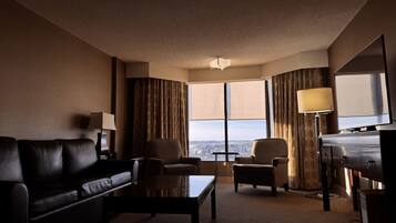 A hotel room with a leather sofa, two armchairs, a coffee table, a TV, and a large window with curtains.