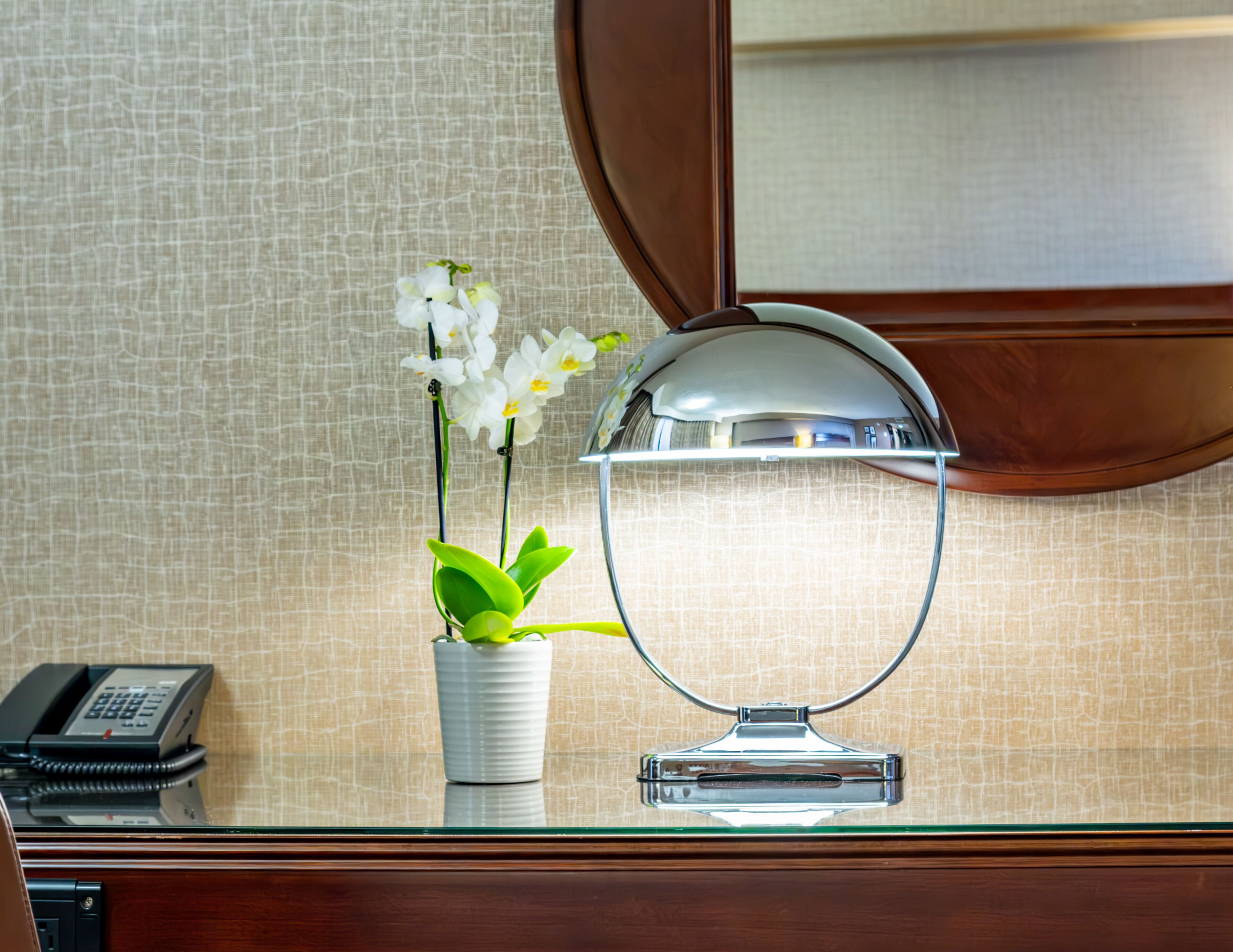 A hotel room with a wooden desk, a telephone, a mirror, and a potted orchid.