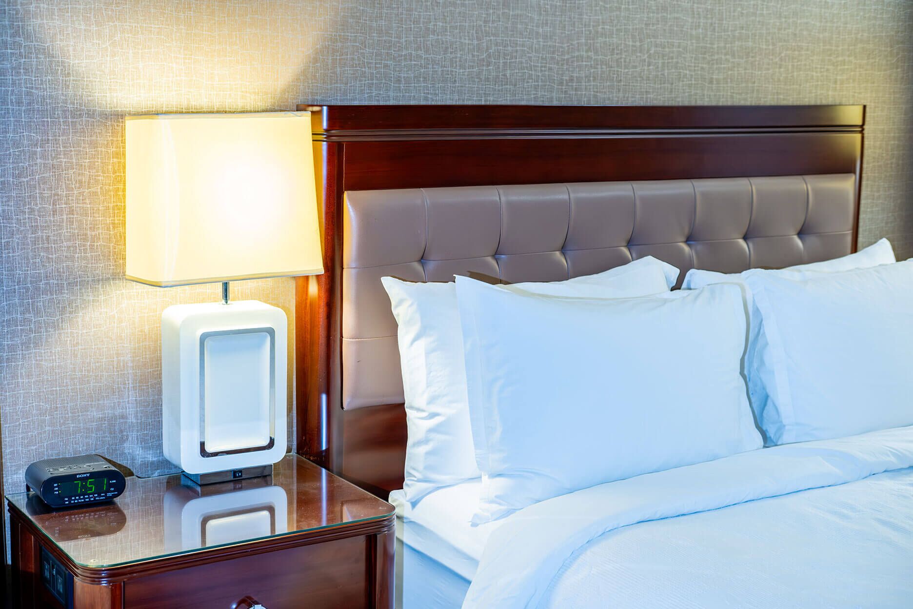 A hotel room with a bed, white linens, a bedside table with a lamp, and a digital clock.