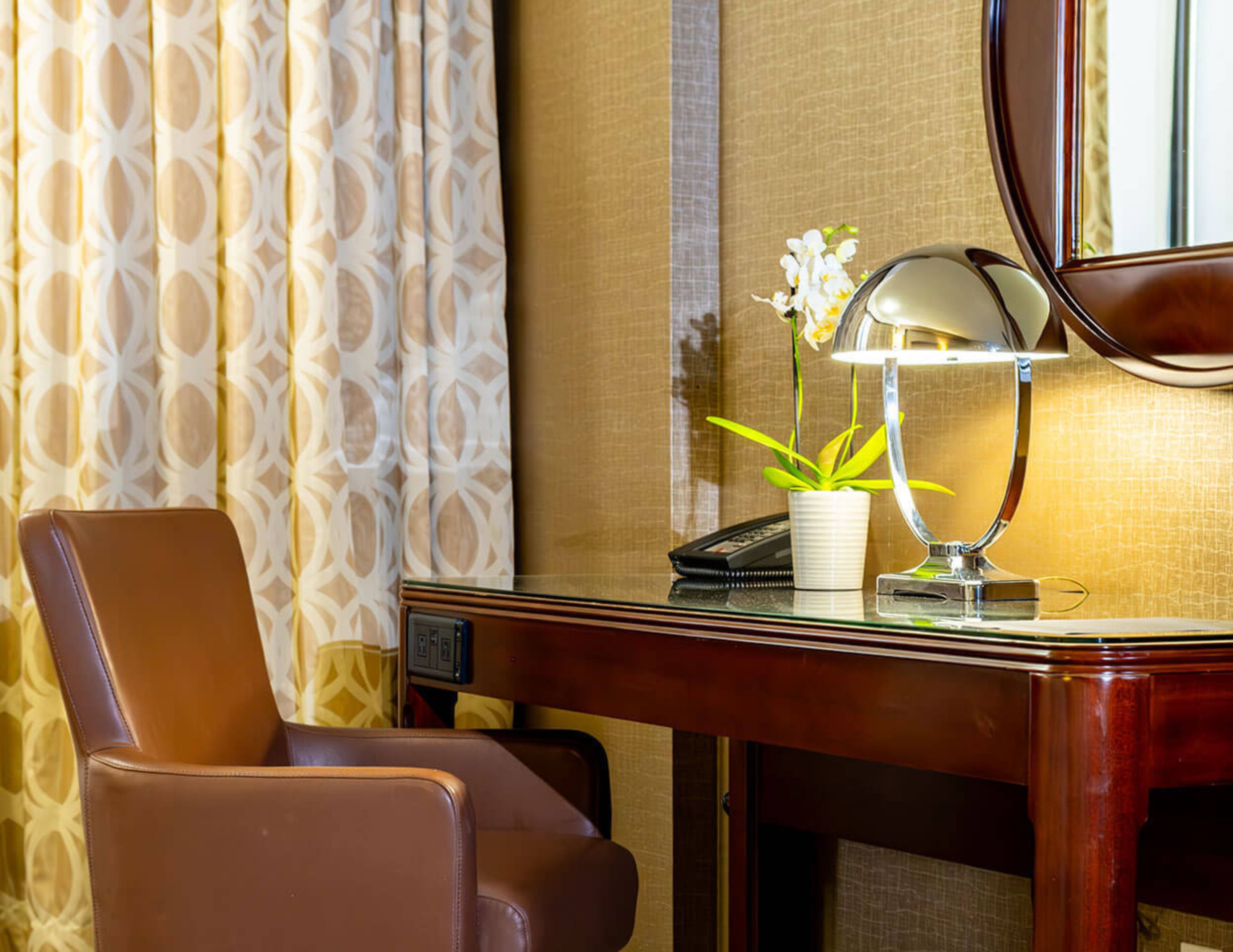 A hotel room with a leather chair, a desk with a lamp, a potted plant, and a telephone.