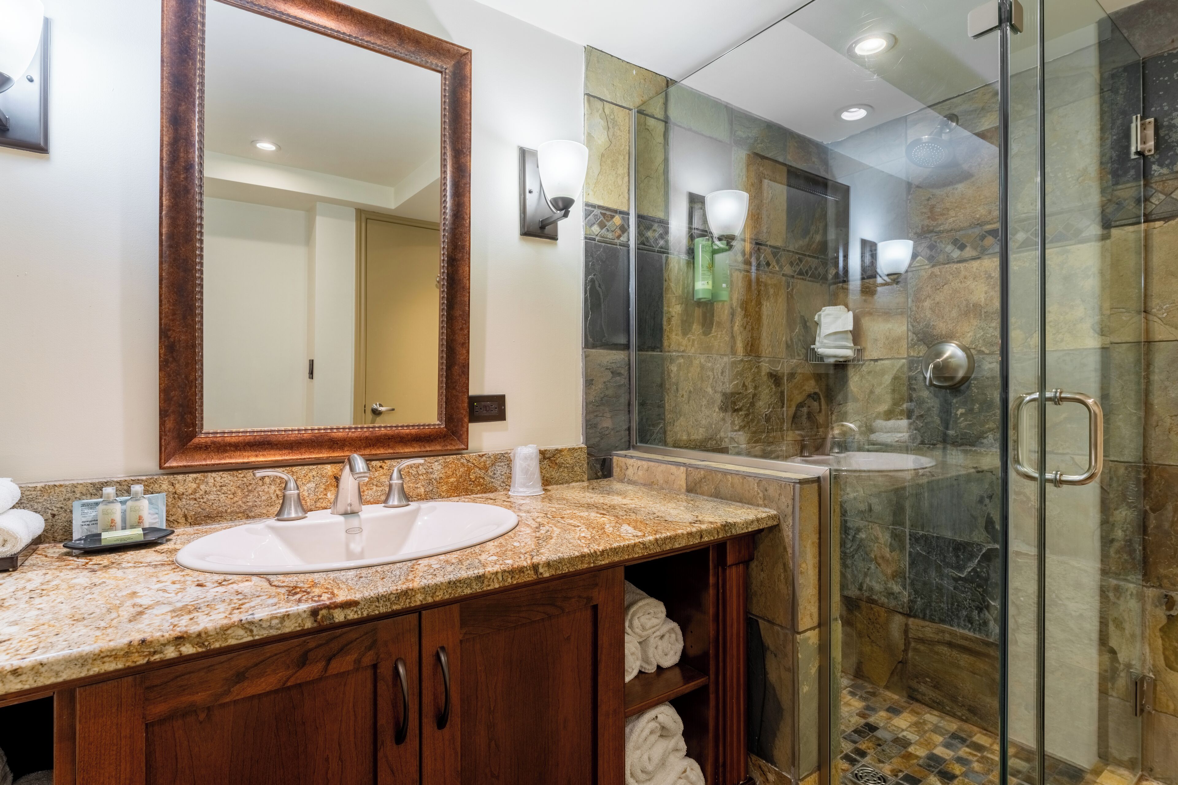 Un baño moderno con encimera de granito, un espejo grande, ducha con cerramiento de vidrio y armarios de madera.