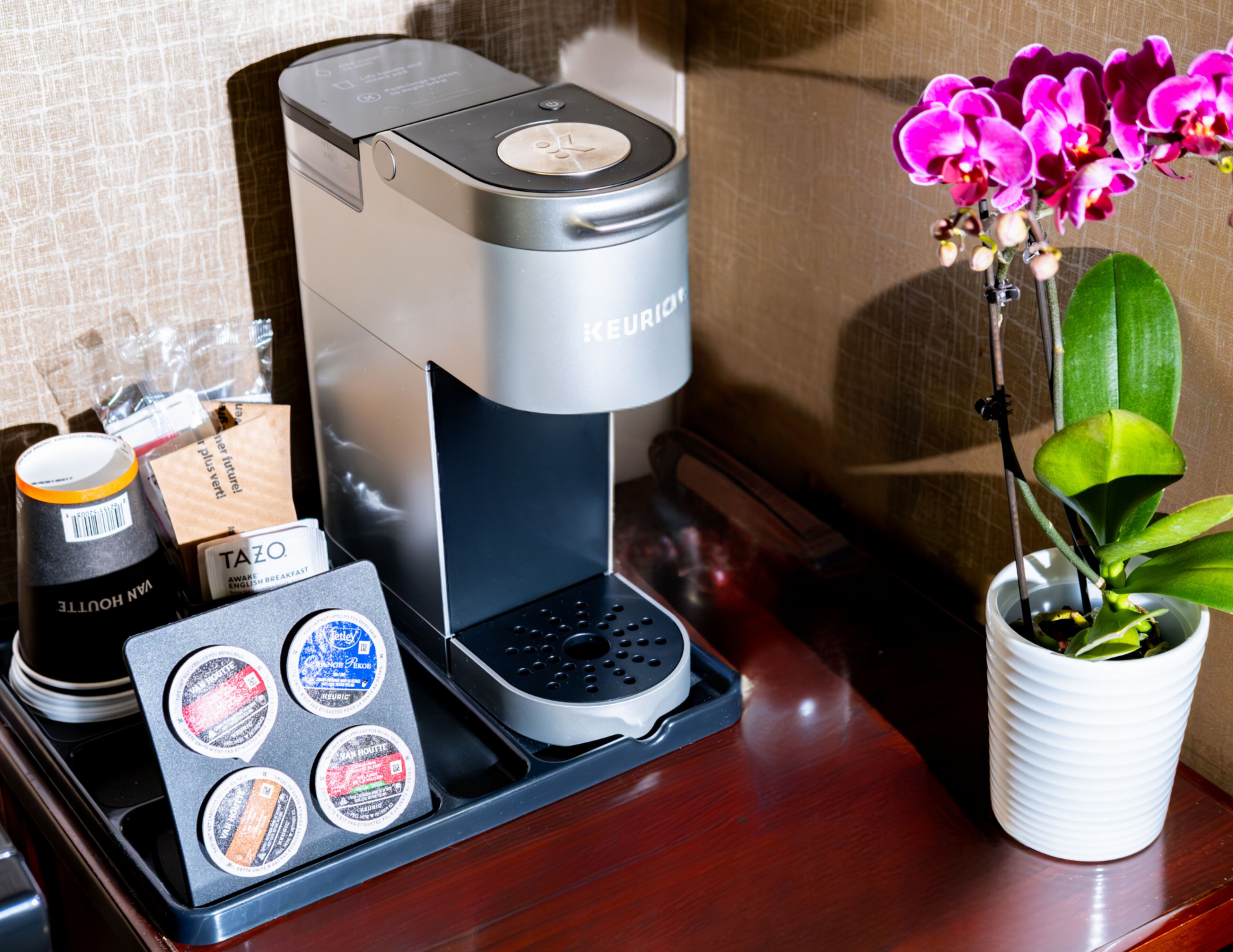 A coffee machine with a tray of coffee pods, a cup of coffee, and a potted orchid on a wooden surface.