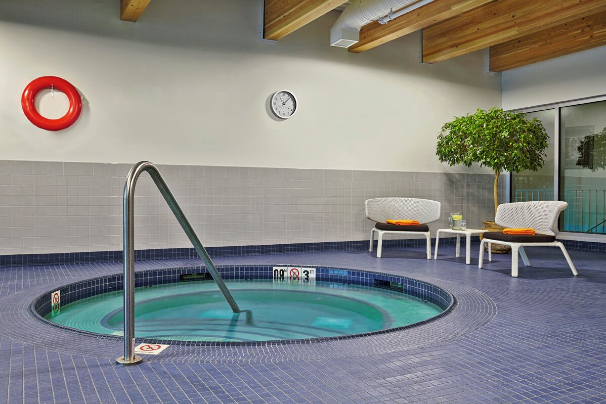 A swimming pool with a metal ladder, surrounded by blue tiles, white lounge chairs, and a red lifebuoy on the wall.