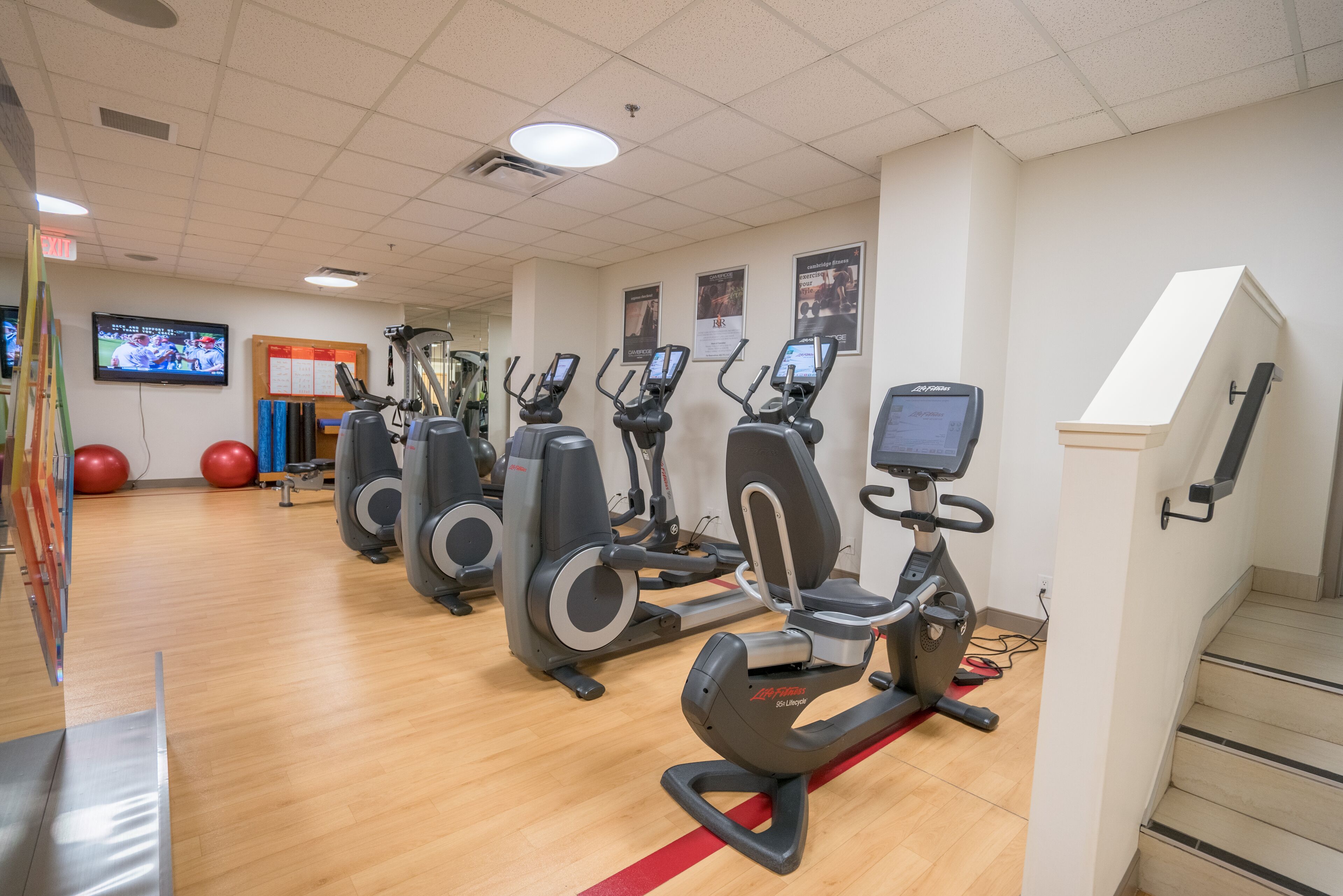 Et fitnesscenter med flere elliptiske træningsmaskiner, røde træningsbolde og et fjernsyn, der viser en sportsbegivenhed.