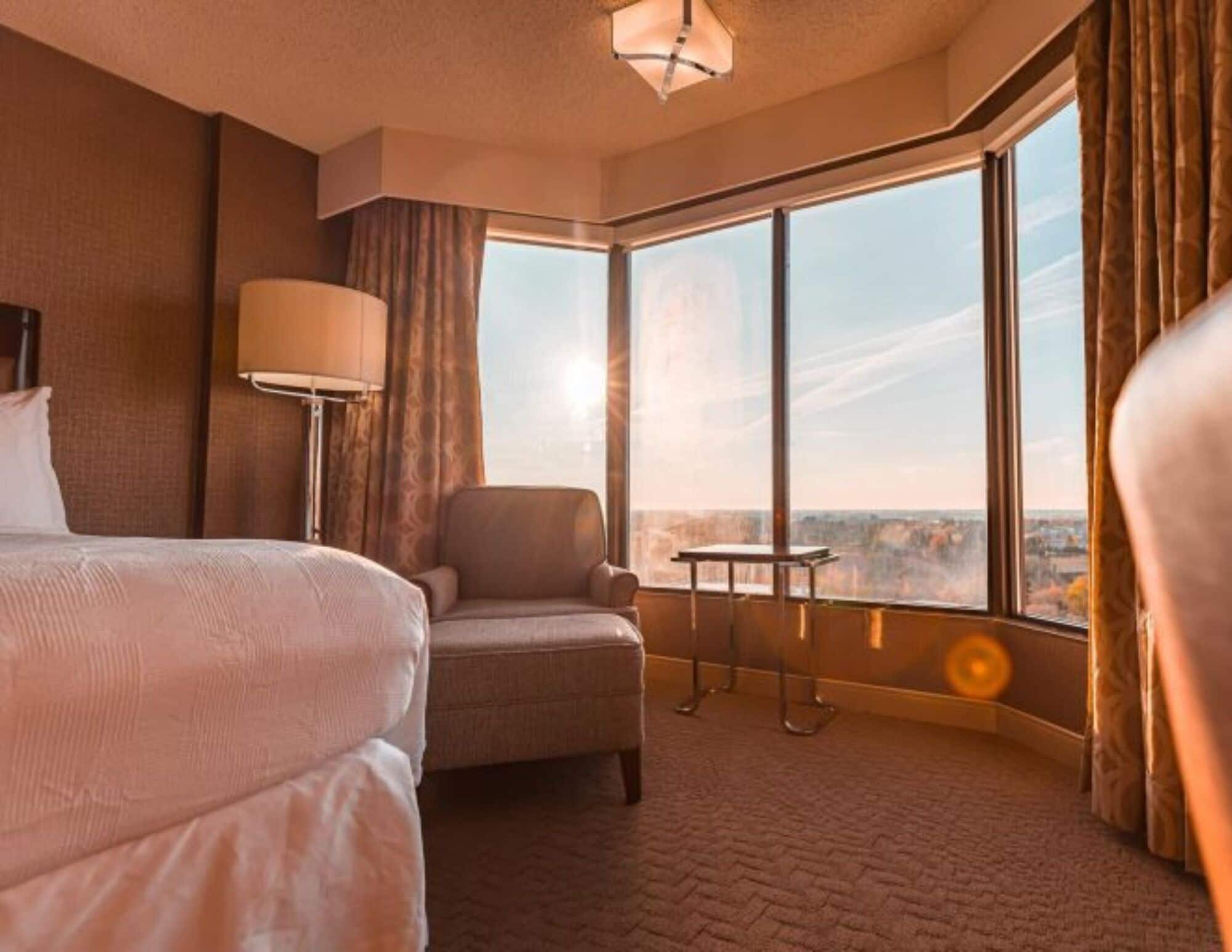 A hotel room with a large bed, a chair, a table, and a view of the city through large windows.