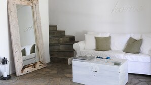 Una stanza con pavimento in pietra, pareti bianche, un divano bianco con cuscini verdi, un settimino bianco e un grande specchio con cornice rustica.