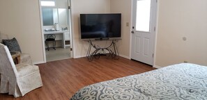 A bedroom with a bed, a television on a stand, a desk with a chair, and a bathroom visible through an open door.