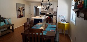 A dining area with a wooden table, chairs, and a yellow side table. There is a kitchen in the background with modern appliances.