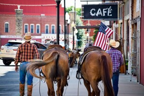 A street scene with horses and riders, a cafe sign, and a building with a red facade.