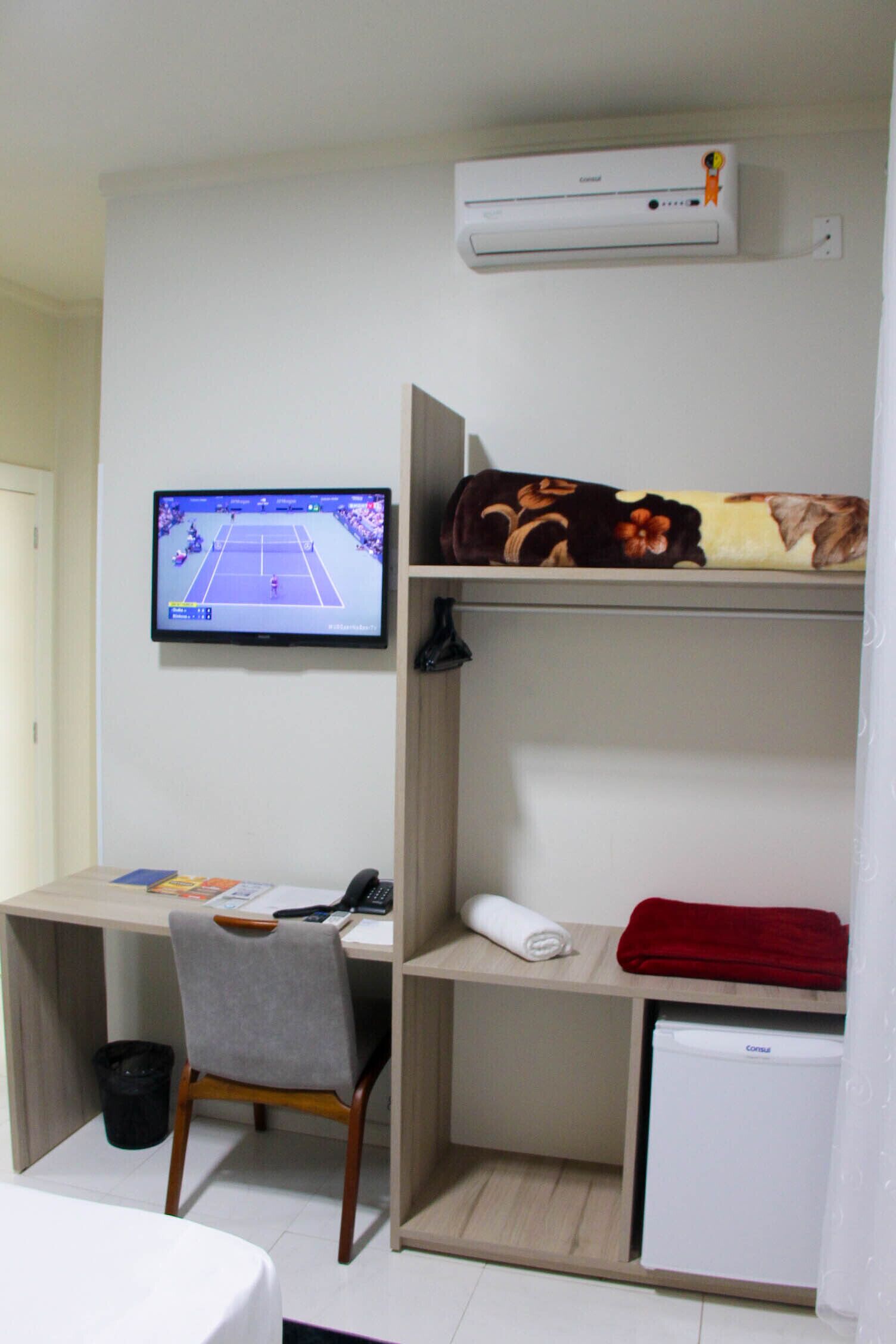 Une chambre de taille modeste équipée d’un bureau, d’une chaise, d’une télévision, d’un climatiseur et d’un petit réfrigérateur.
