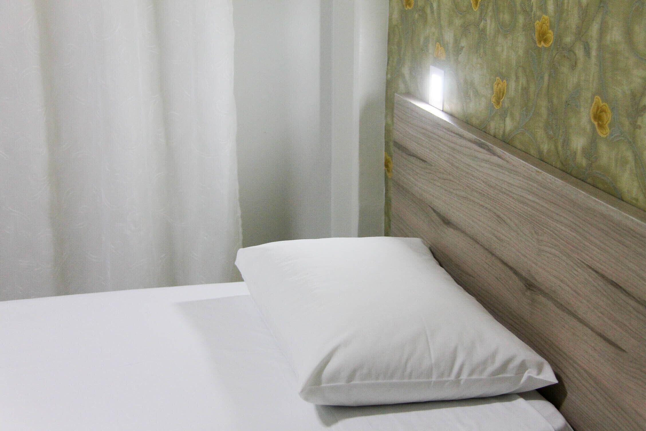 Una cama con ropa blanca y una única almohada blanca, cabecera de madera y pared con estampado floral.