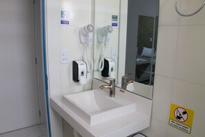 A bathroom with a white sink, a mirror, two hair dryers, and a no-paper sign on the wall.