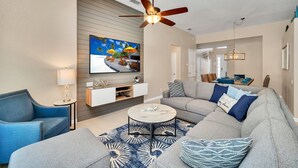 Una sala de estar moderna con un sofá gris, un sillón azul y una mesa de centro. Se ve un ventilador de techo y un televisor montado en la pared.