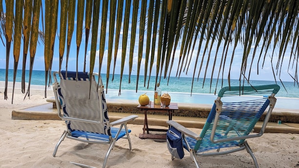 Dues cadires de platja amb una taula al mig, una ampolla i un coco a la sorra prop de l'oceà.