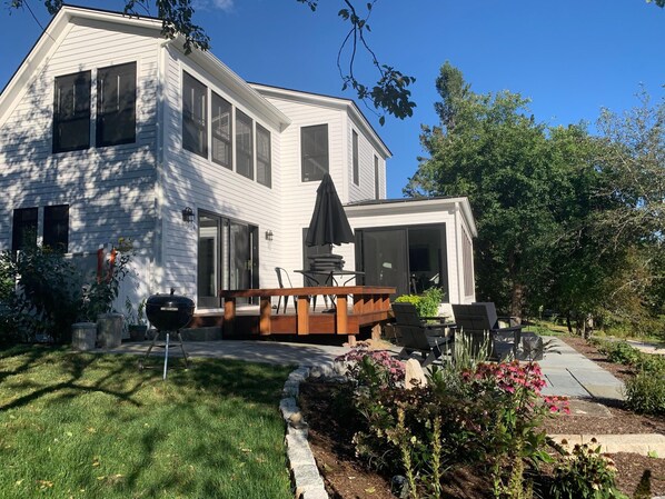 A modern two-story house with a patio area featuring a grill, outdoor furniture, and a garden with various plants and flowers.