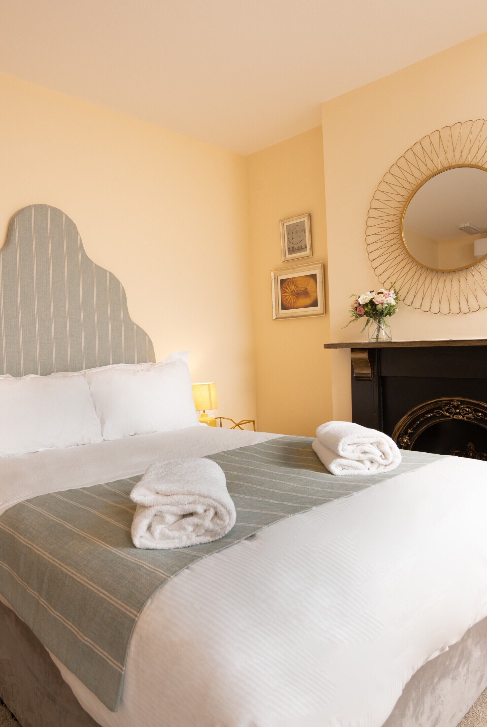 A neatly made bed with pillows and a folded towel, a bedside lamp, a framed picture, and a mirror on the wall.