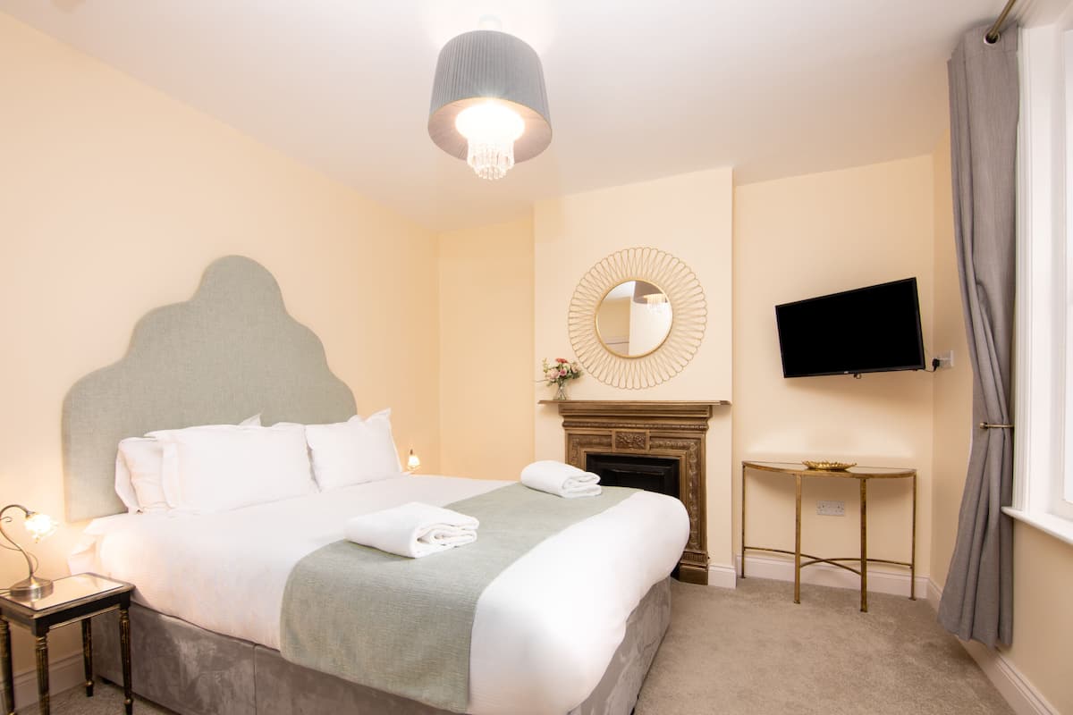 A hotel room with a large bed, a television mounted on the wall, a small table, and a fireplace.