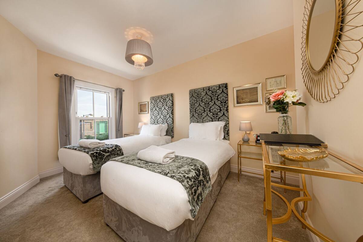 A hotel room with two beds, a desk, a mirror, and a chandelier.