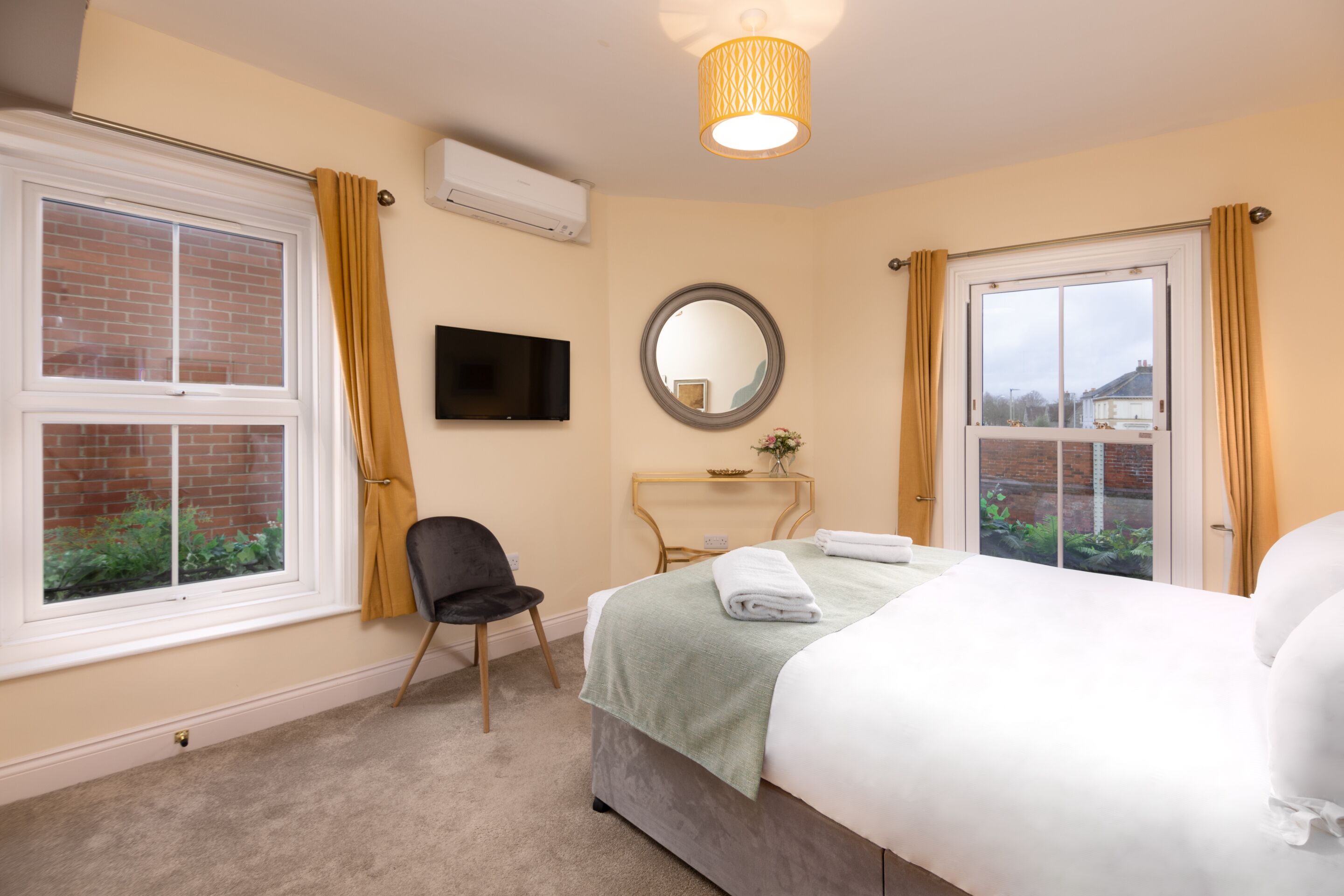 A hotel room with a bed, a chair, a TV, a mirror, and windows with curtains.