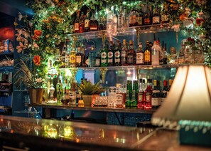 Una zona de bar con estantes llenos de diferentes botellas de licor, plantas decorativas y un expositor de vidrio.