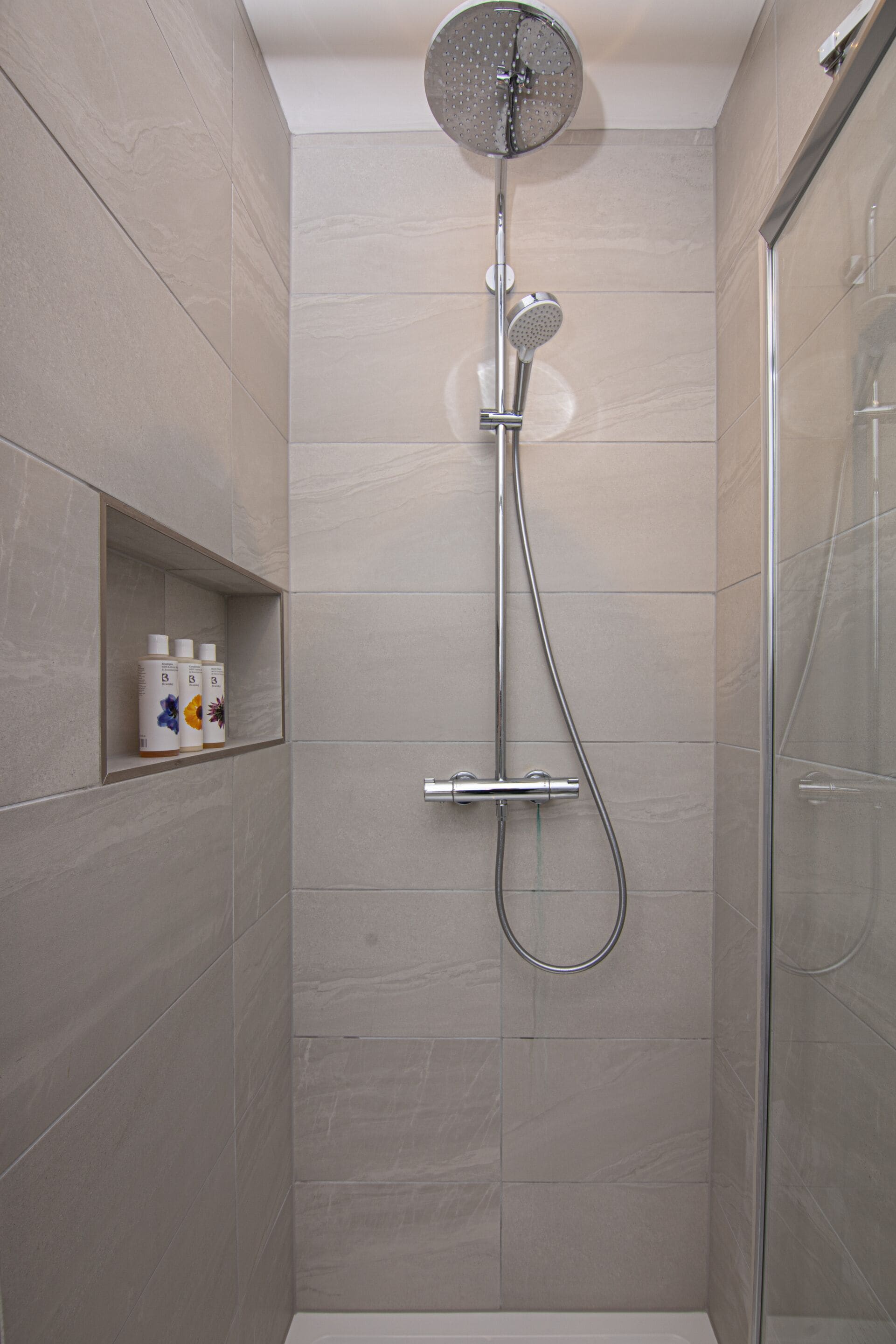 A modern shower with a rain showerhead and handheld showerhead, a glass door, and a shelf with toiletries.