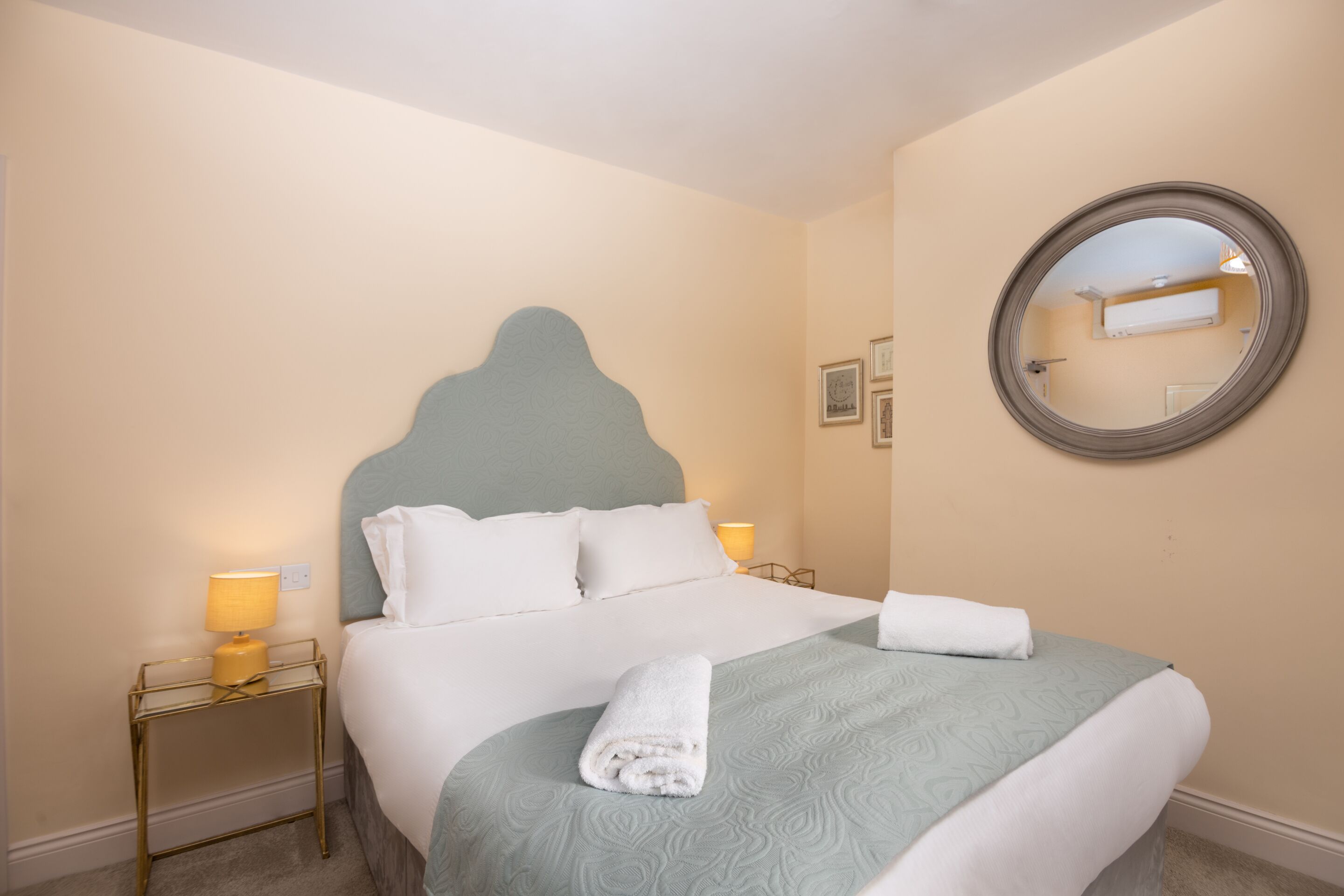 A neatly made bed with a light blue headboard, a bedside table with a lamp, and a round mirror on the wall.