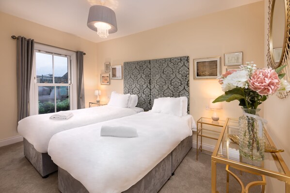 A hotel room with two beds, a large window with curtains, a chandelier, framed artwork, and a vase with flowers.