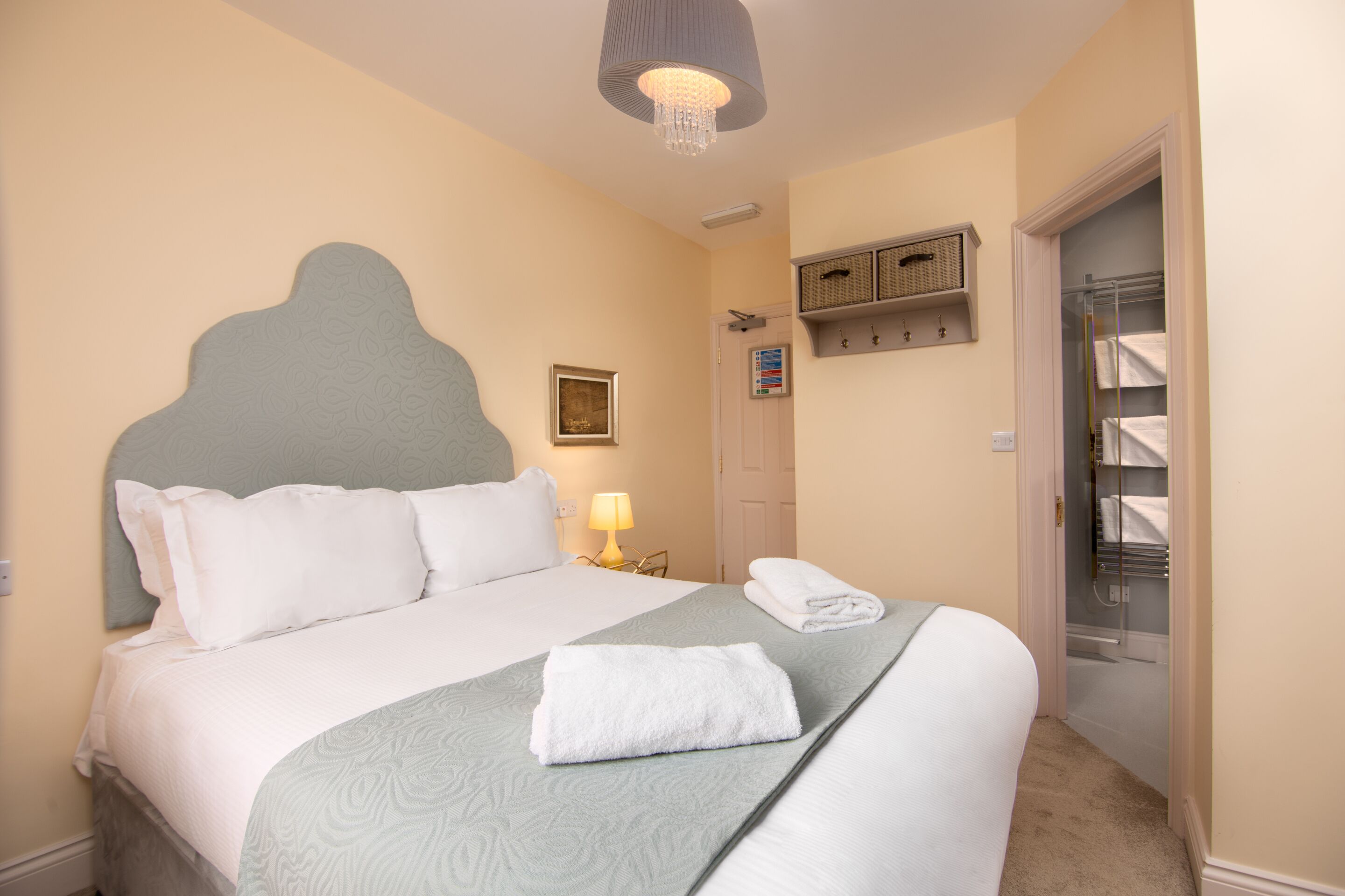 A neatly made bed with white linens and a folded towel, a bedside lamp, a framed picture on the wall, and a storage shelf above a door.