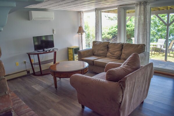 A living room with a sofa, a round coffee table, a television on a stand, and a window with a view of greenery outside. - Amazing pond views and unforgettable sunsets from all levels! (Dennis Port)