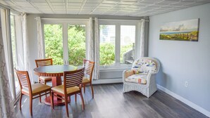 Ruokailutila, jossa on pyöreä pöytä ja tuolit, rottinkituoli sekä suuret ikkunat, joista avautuu näkymä vehreään maisemaan.