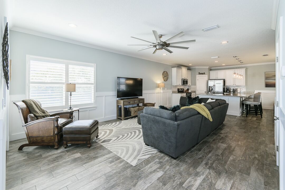 A spacious living room with a gray sofa, a wooden coffee table, and a TV. The kitchen is visible in the background with white cabinets and a dining area.