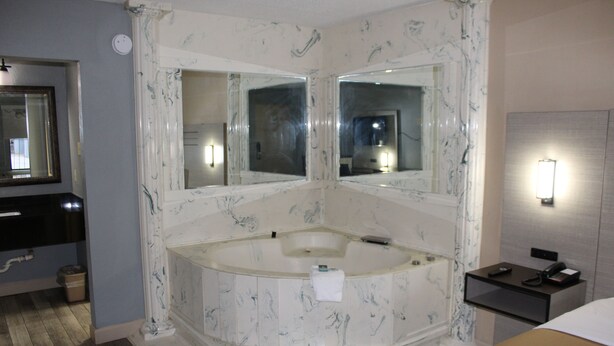 Une salle de bain avec une baignoire en marbre, deux miroirs fixés au mur, un porte-serviettes et une table de chevet avec un téléphone et une lampe.